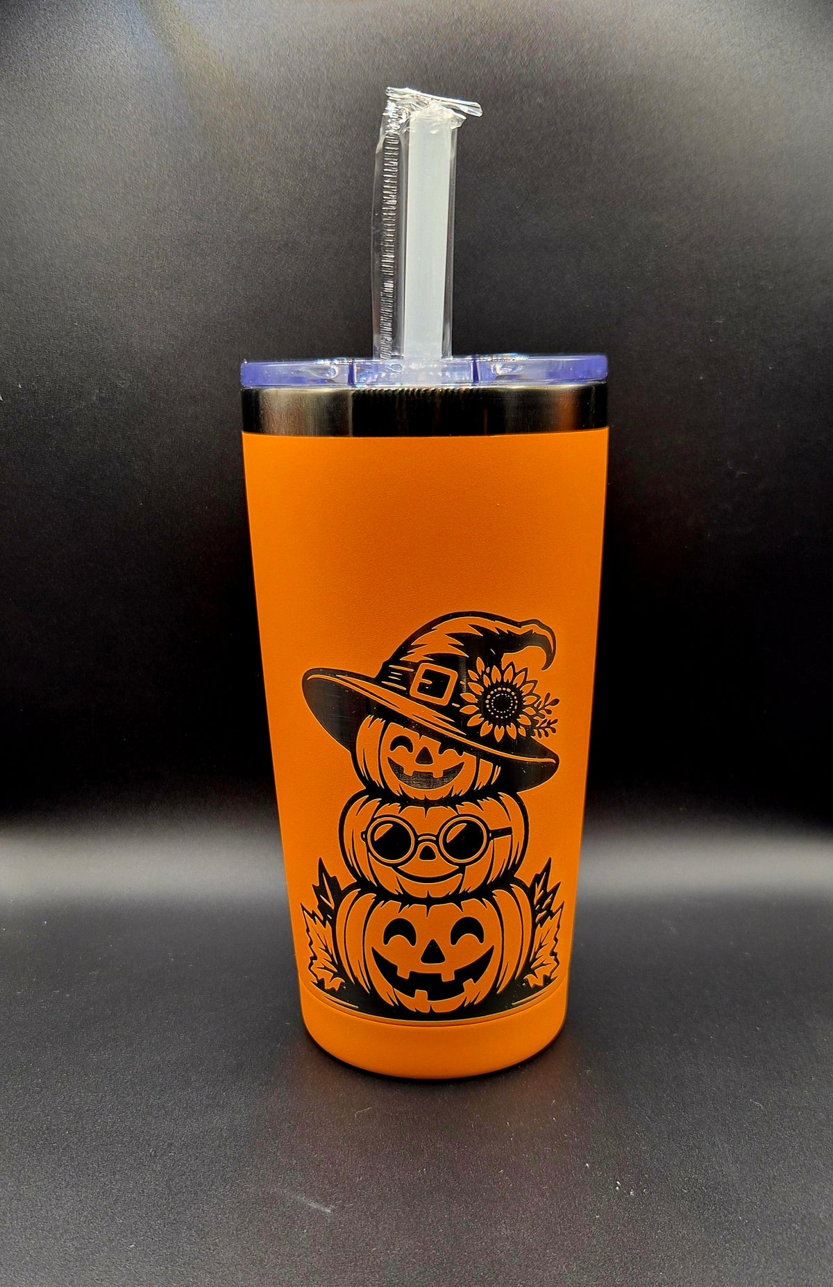 Bright orange 20 oz stainless steel Halloween tumbler with black laser-engraved artwork of three stacked smiling jack-o'-lantern pumpkins, round glasses, autumn leaves, and a witch hat decorated with a sunflower. Clear lid and straw included.