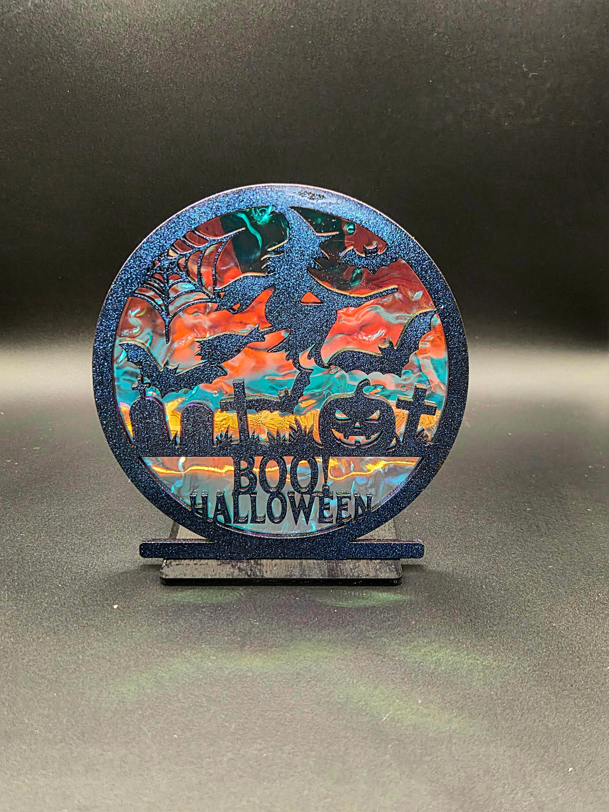 Round Halloween decoration featuring bats, gravestones, jack-o'-lantern and BOO HALLOWEEN lettering over an iridescent background.
