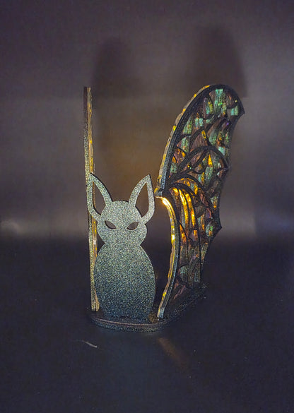 Black bat silhouette between glowing iridescent acrylic wings, illuminated by an LED tealight in a dark Halloween display.