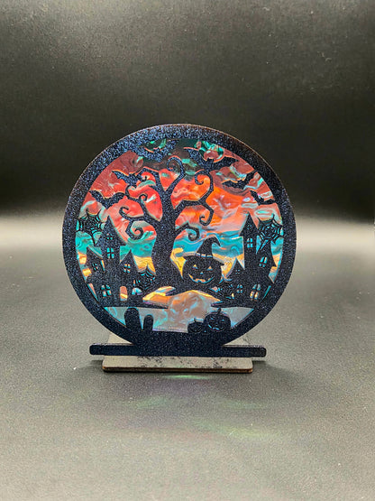 Freestanding Halloween decoration with black haunted houses, twisted tree, bats and pumpkins against colorful iridescent backing.