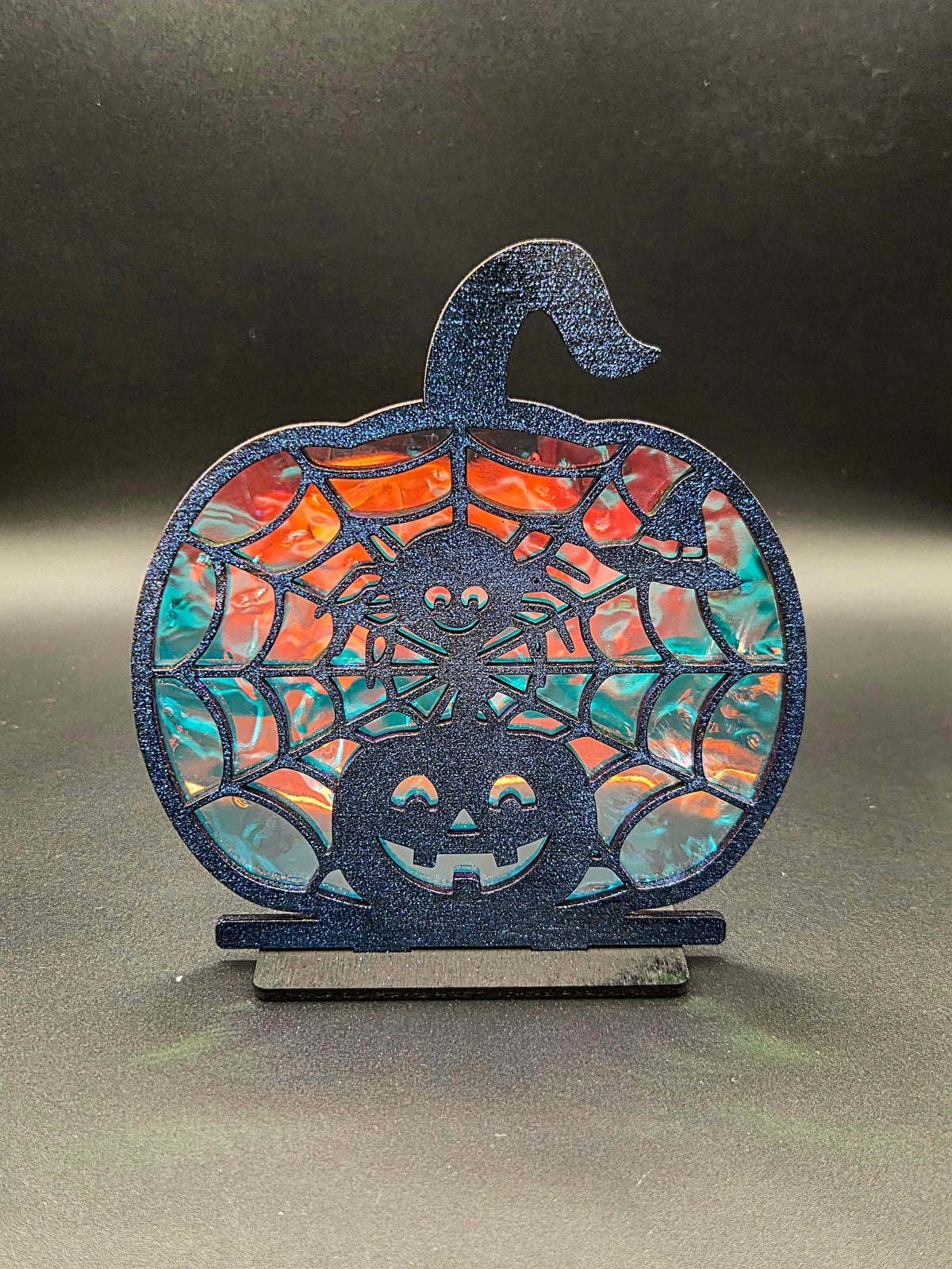 Pumpkin-shaped Halloween decoration with smiling spider, spiderwebs and jack-o'-lantern against orange and turquoise iridescent backing.